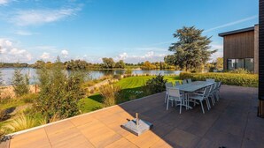 Eine Terrasse mit Tisch und Stühlen mit Blick auf einen See, umgeben von viel Grün und einem modernen Gebäude.
