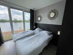 A bedroom with a bed, a large window, and two wall mirrors. - Aan de Maas - Maas Villa Waterzicht | 8p (Kerkdriel)