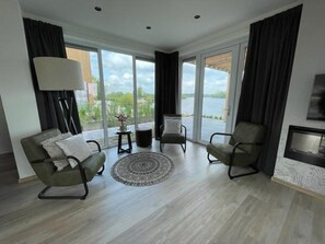 A modern living room with a sofa, armchairs, a coffee table, and a fireplace. Large windows offer a view of a body of water. - Aan de Maas - Maas Villa Waterzicht | 8p (Kerkdriel)