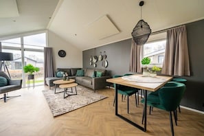 Ein modernes Wohnzimmer mit Sofa, Couchtisch, Esstisch und Stühlen. Es gibt ein großes Fenster mit Vorhängen, eine Pendelleuchte und eine wandmontierte Uhr.