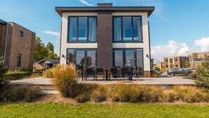 En modern byggnad i två våningar med stora fönster, en uteplats med utomhusmöbler och en välskött gräsmatta.