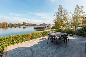 A patio with a table and chairs overlooking a calm lake, with boats docked nearby. - Aan de Maas - Gelderse Lande Waterzicht | 6p (Kerkdriel)