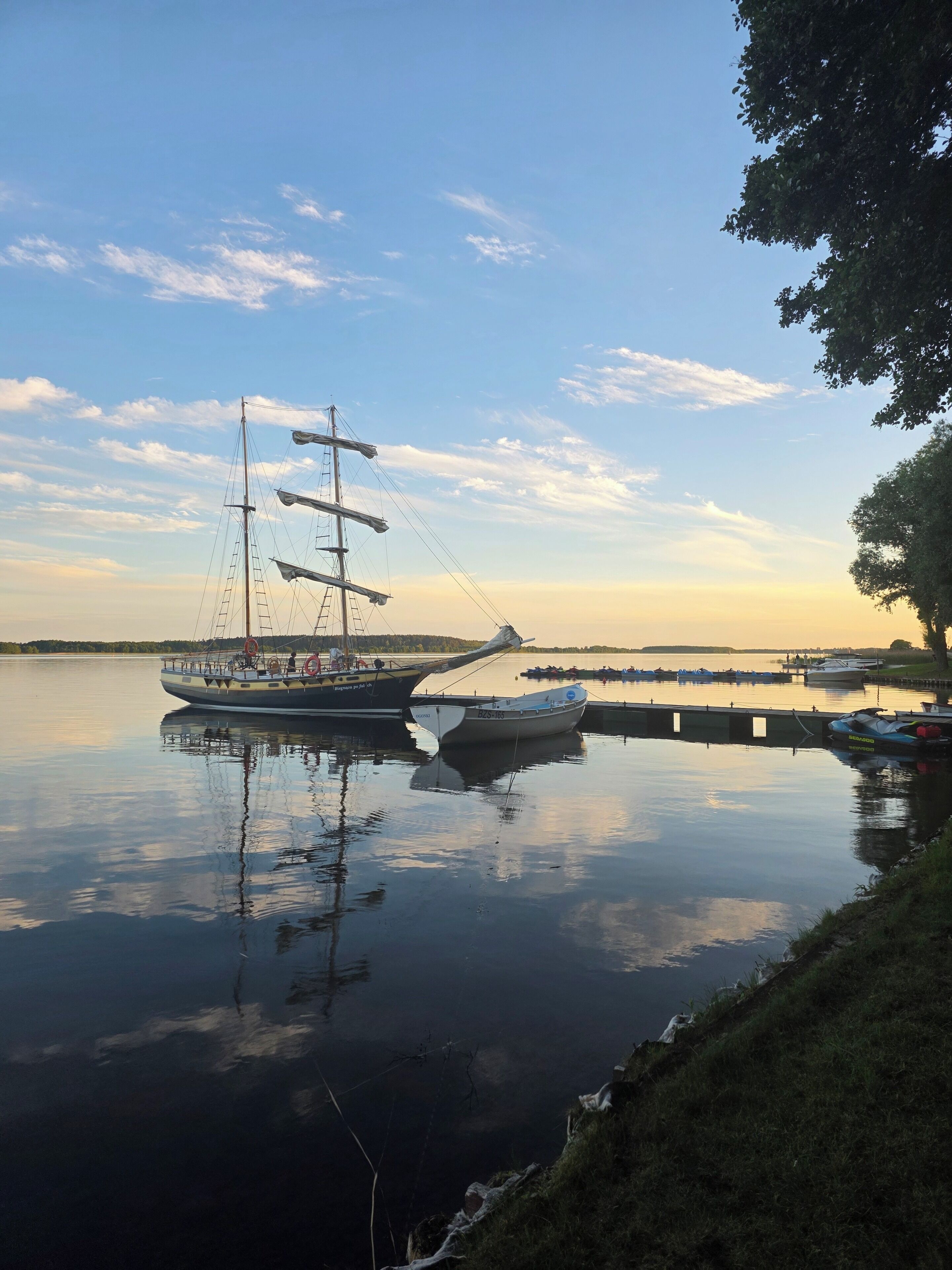 Un lago tranquilo con un barco de vela amarrado en un muelle, rodeado de aguas serenas y un cielo despejado.