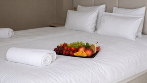 A neatly made bed with white linens and pillows, a tray of assorted fruits, and rolled towels.