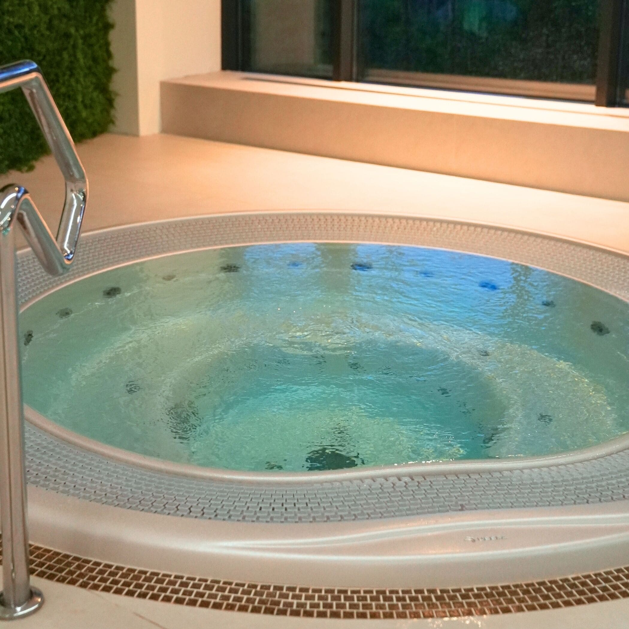 Een hottub met een helder wateroppervlak, omgeven door een betegelde rand, en een metalen trap aan de linkerkant.