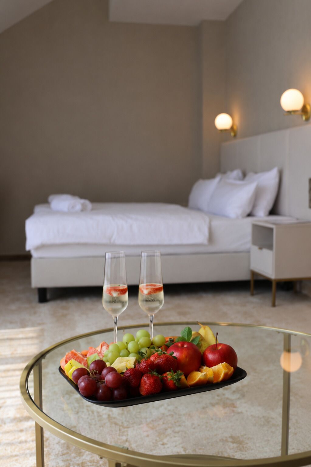 Quarto de hotel com cama, mesa de cabeceira e uma mesinha de centro com frutas e duas taças de vinho branco.