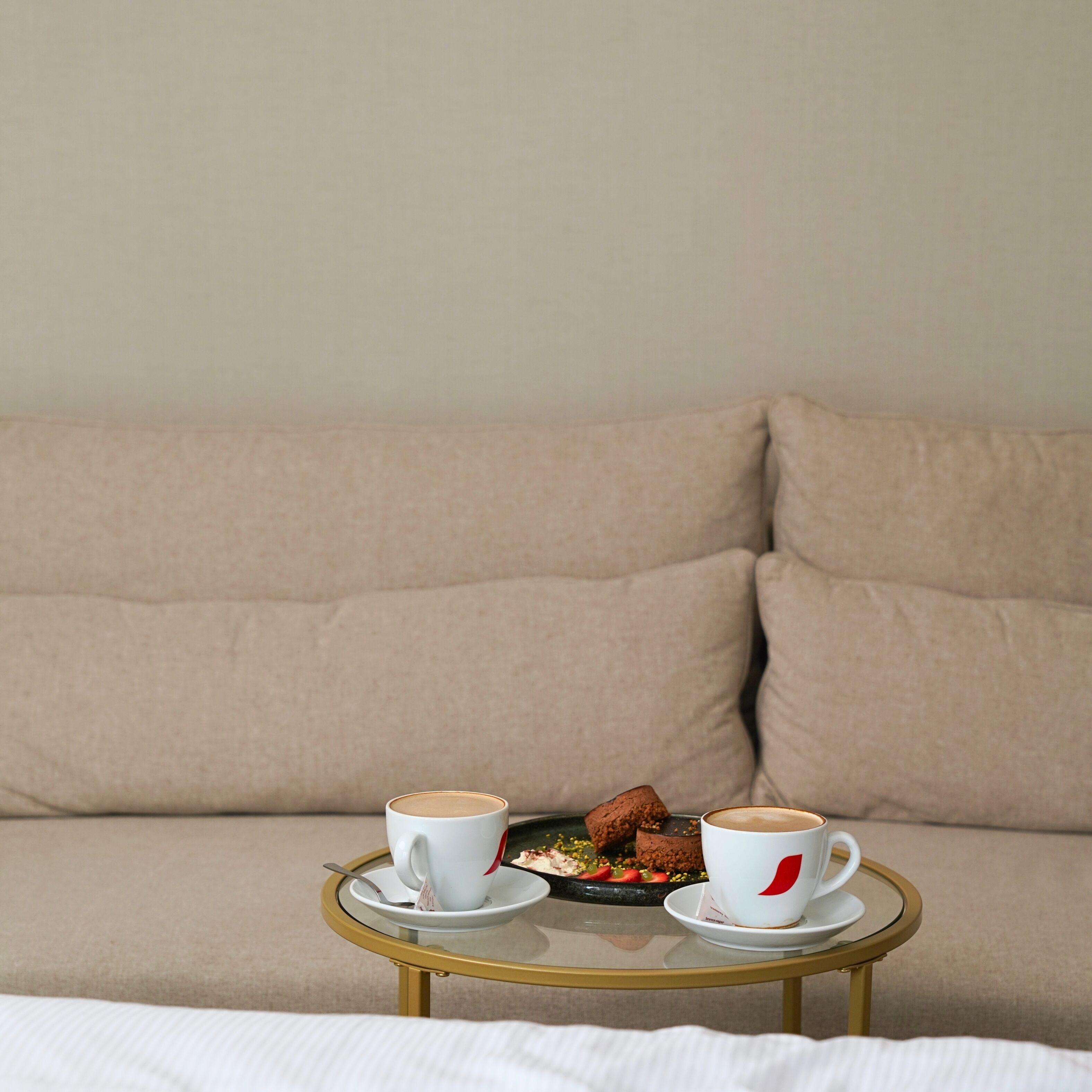 A bed with a white comforter, a round glass table with two white cups and a plate of food, and a beige sofa in the background.