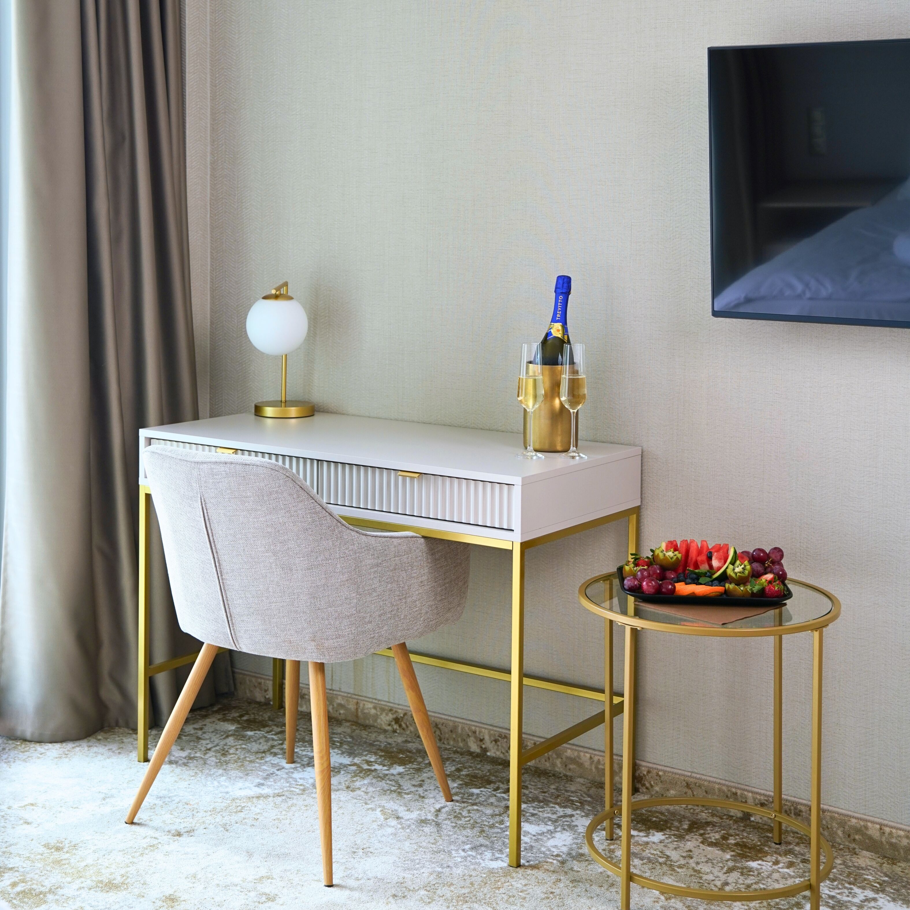Una habitación moderna con un escritorio blanco, una silla con marco dorado, una pequeña mesa redonda con una bandeja de frutas y una botella de champán.