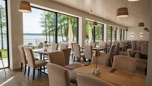 A spacious dining area with multiple tables and chairs, large windows with a view of trees and water, and pendant lighting.
