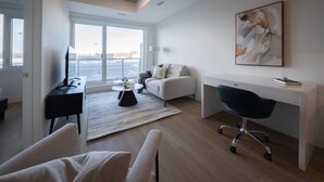 Ein modernes Wohnzimmer mit großem Fenster, einem weißen Sofa, einem schwarzen Bürostuhl, einem weißen Schreibtisch und einem Gemälde an der Wand.