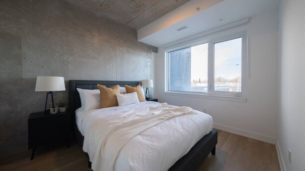 Ein Schlafzimmer mit einem großen Bett, einem Nachttisch mit Lampe, einem Fenster mit Blick und einer Betondecke.