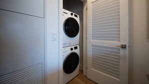 Ein Waschraum mit zwei übereinander gestapelten Waschmaschinen und Trocknern, einer Tür mit horizontalen Lamellen und einem Lichtschalter an der Wand.