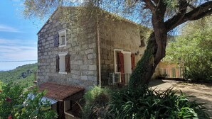 A stone building with a small porch, a tree in front, and a view of hills in the background. - Stone village house in the heart of nature and 15 minutes from the beaches (Conca)
