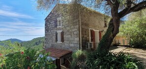 A stone building with a small porch, a tree in front, and a view of hills in the background. - Stone village house in the heart of nature and 15 minutes from the beaches (Conca)