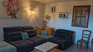 A living room with a sofa, ottoman, coffee table, wall art, and a window with a wooden frame. - Stone village house in the heart of nature and 15 minutes from the beaches (Conca)