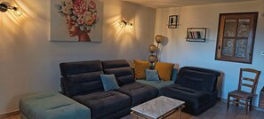 A living room with a sofa, ottoman, coffee table, wall art, and a window with a wooden frame. - Stone village house in the heart of nature and 15 minutes from the beaches (Conca)