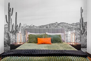 Ein Schlafzimmer mit einem großen Bett, einem grau gepolsterten Kopfteil und einer Wandmalerei zum Thema Wüste.