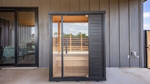 Entrada de un edificio moderno con puerta corredera de vidrio, terraza de madera y vista a un área exterior cercada.
