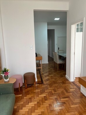 Ein Zimmer mit Fischgrätparkett, einem grünen Sofa, einem kleinen runden Tisch mit einer Pflanze und einem rosa Hocker.