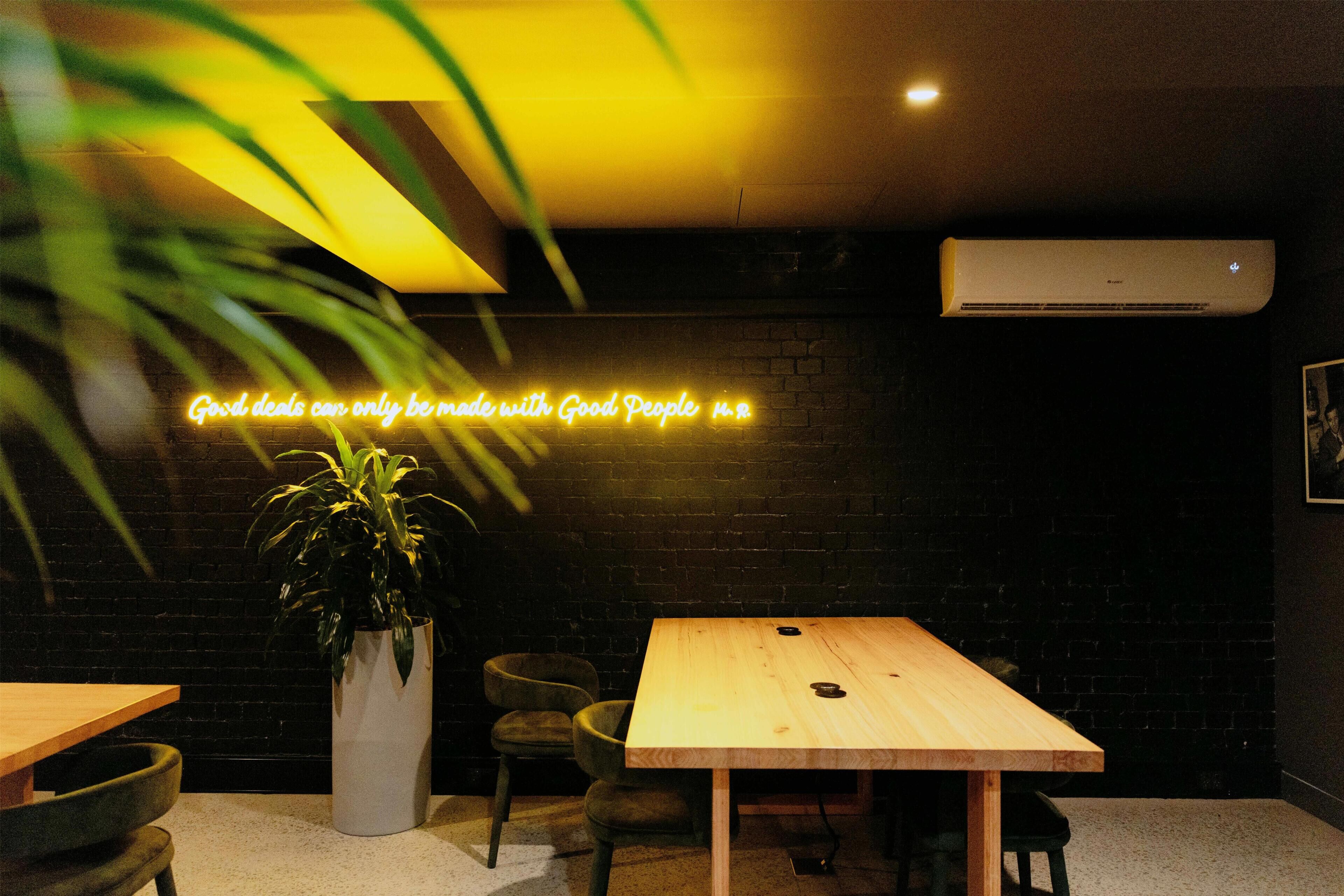 A room with a neon sign, a potted plant, a table, and chairs.