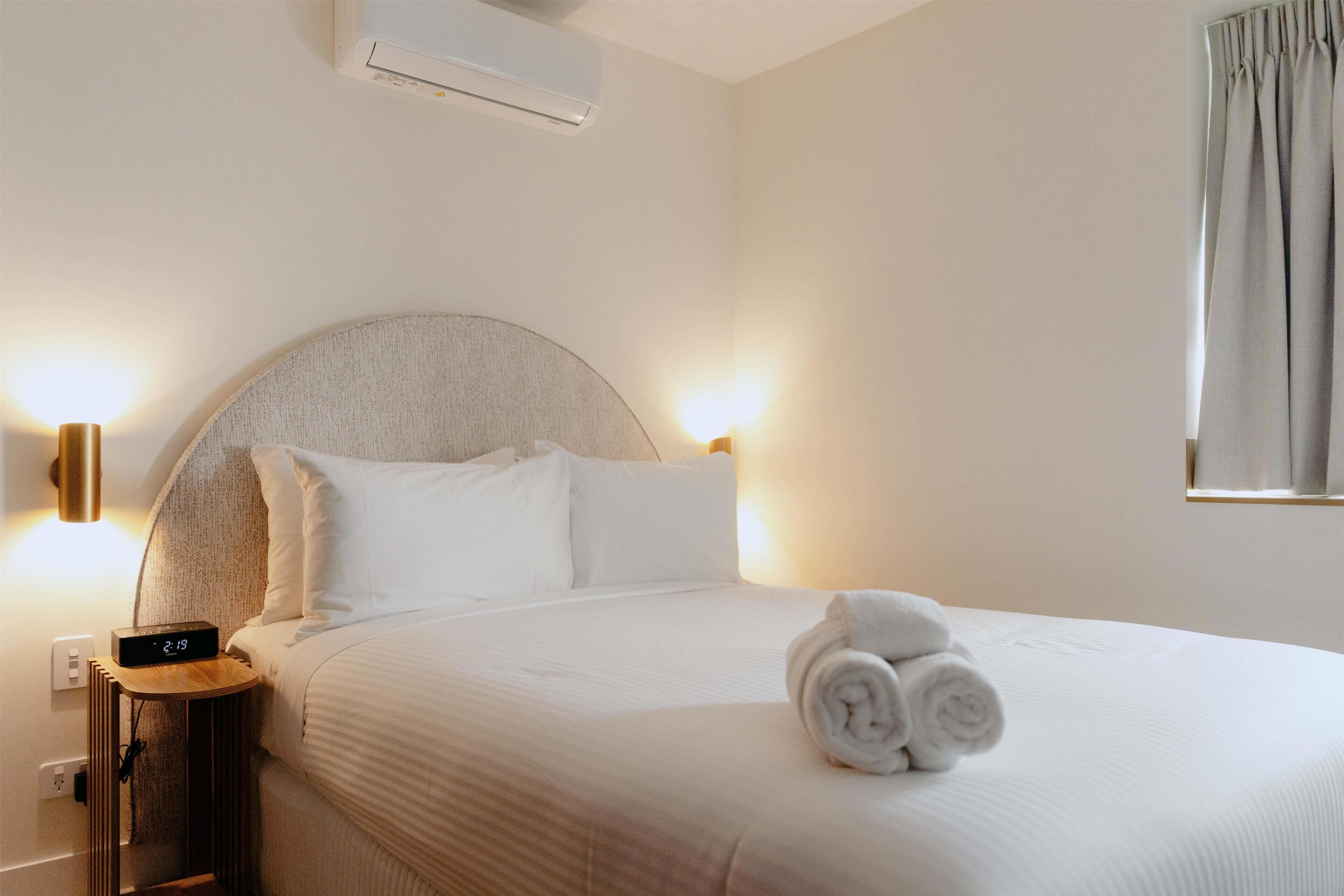 A hotel room with a bed, a bedside table with a clock, an air conditioner, and a window with curtains.