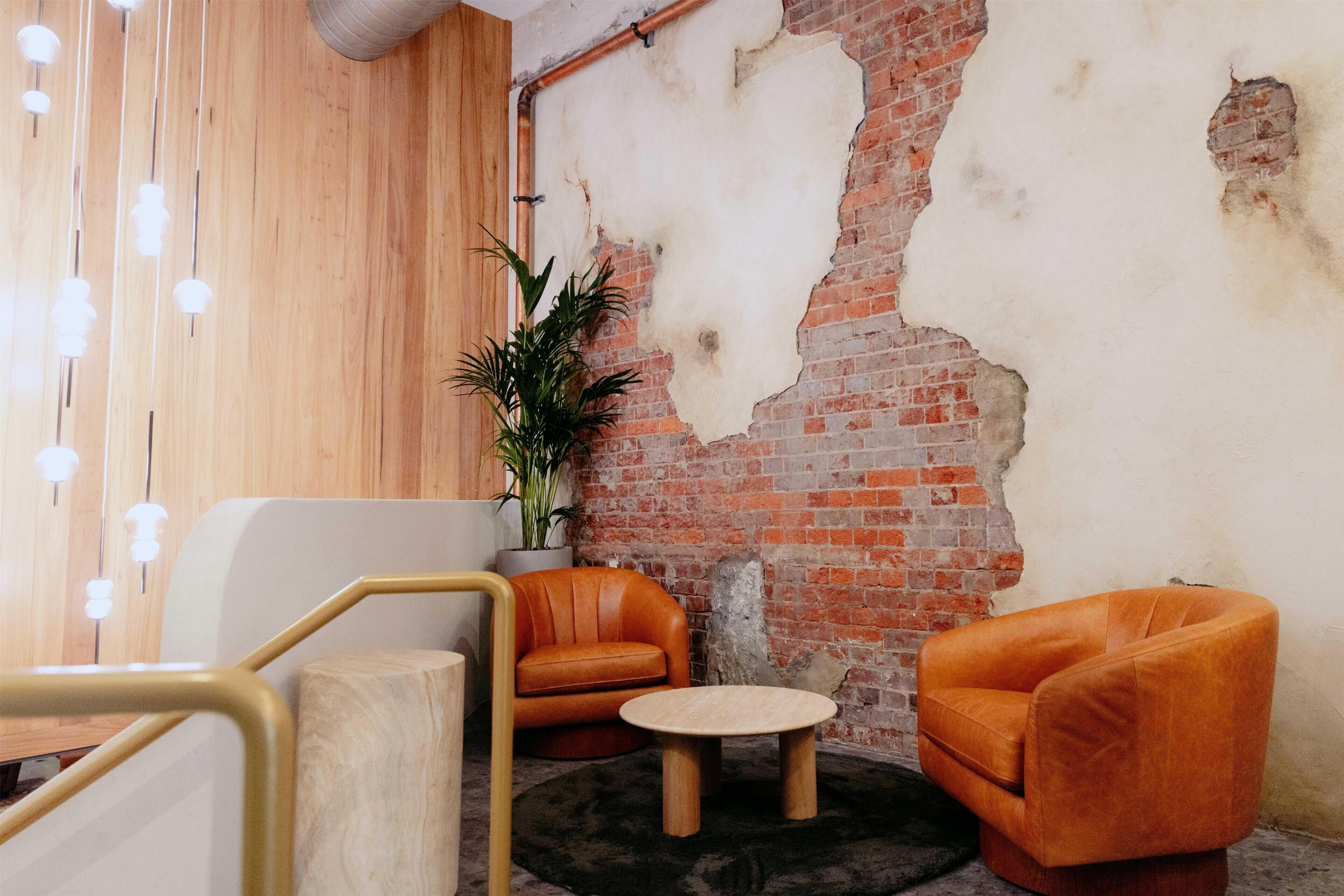 A room with a brick wall, a wooden floor, a round table, two orange chairs, a potted plant, and a staircase with a wooden railing.