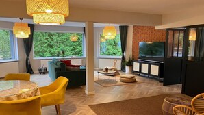 En moderne stue med spiseplads, store vinduer og et fjernsyn.