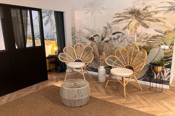 A room with a large mural, wicker chairs, a round ottoman, and a small table with a lamp.
