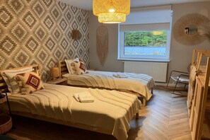 A bedroom with two beds, a large window, a hanging light fixture, and a wall-mounted dreamcatcher.