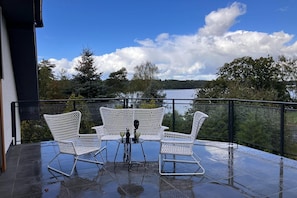 Un balcon avec deux fauteuils en osier blancs et une petite table, donnant sur un lac bordé d’arbres et sous un ciel dégagé.