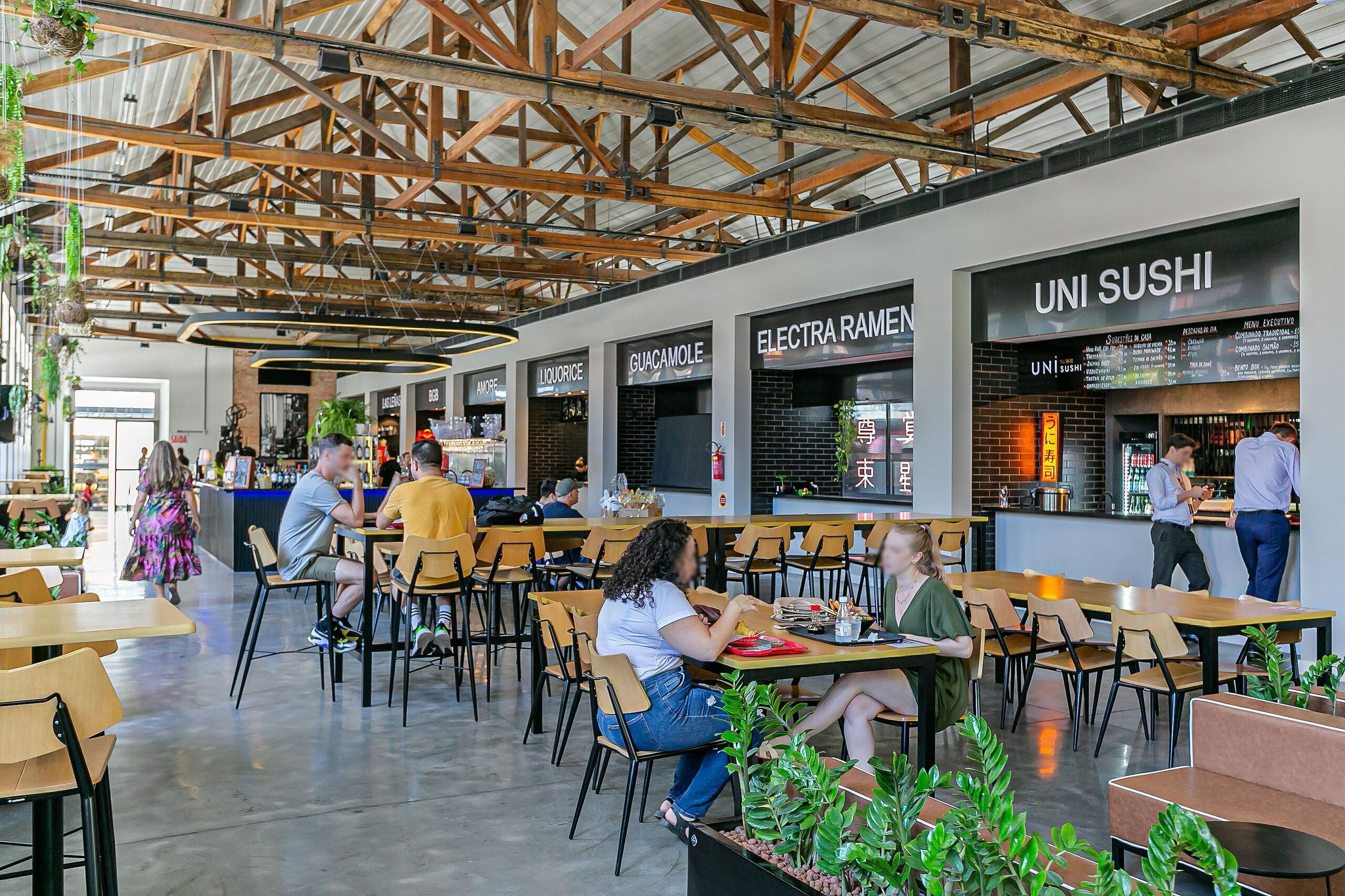 Ein moderner Essbereich mit verschiedenen Sitzgelegenheiten, darunter Tische und Stühle. Mehrere Restaurantschilder sind vorhanden, darunter eines für 'UNI SUSHI'. Der Raum zeichnet sich durch eine hohe Decke mit freiliegenden Holzbalken und hängenden Pflanzen aus.