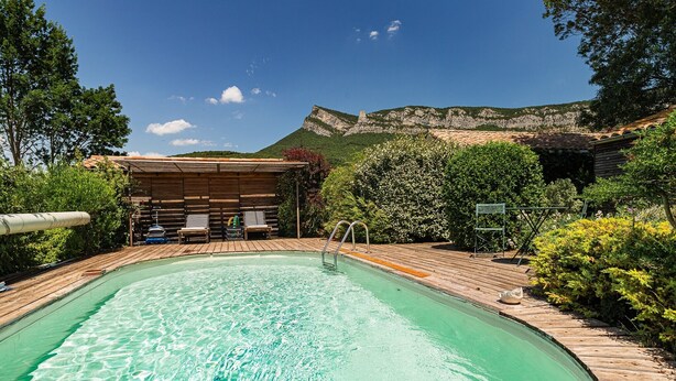 Una piscina con terrazza in legno e poltrone prendisole, immersa nel verde con montagne sullo sfondo.