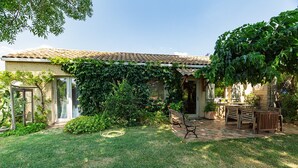 Una casa con tetto in tegole, un patio coperto con mobili in legno e un giardino con prato e siepi.