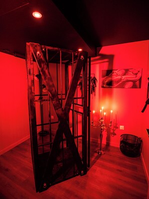 A room with a red-lit cage-like structure, a candelabra, and a framed picture on the wall. - Nuit Défendue - Sauna Jacuzzi Secret room (Tournus)