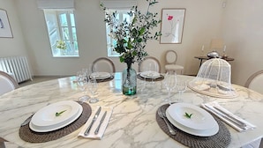 Una mesa redonda de comedor con platos blancos, vasos transparentes y un jarrón verde con ramas.
