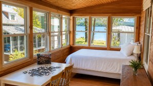A cozy cabin with a bed, a table with a puzzle, and large windows overlooking a lake. - Sunapee Shore and Summit (Newbury)