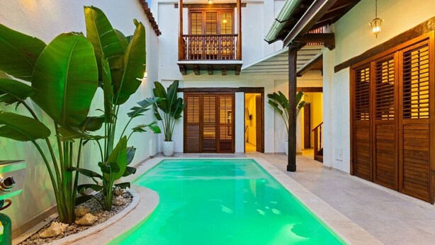 Ein Pool mit geschwungener Kante, umgeben von üppigen Grünpflanzen und einem zweistöckigen Gebäude mit Holztüren und einem Balkon.