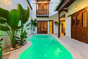 A swimming pool with a curved edge, surrounded by large green plants and a two-story building with wooden doors and a balcony. - Casa Sol - Villa in Getsemaní with Large Pool (Cartagena)