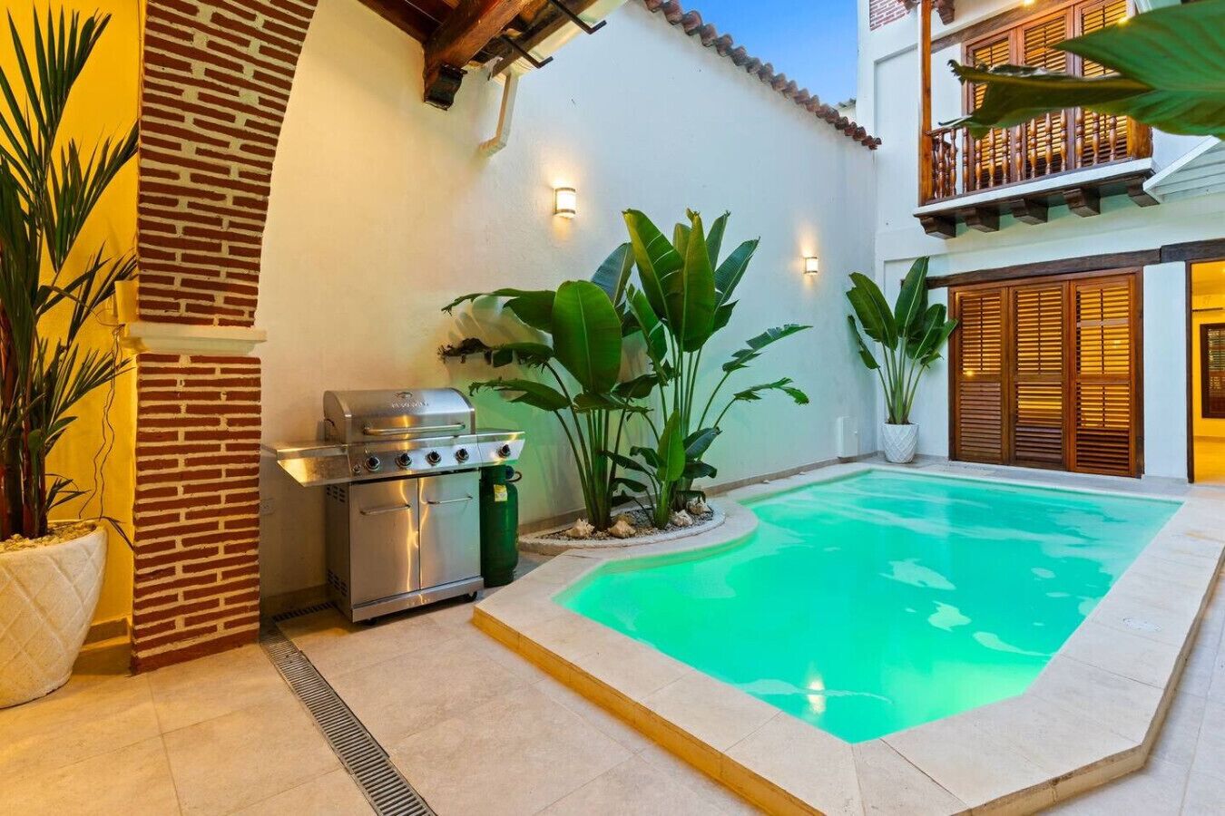 A poolside area with a barbecue grill, potted plants, and a balcony with wooden shutters.