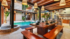 Ein Essbereich am Pool mit Holzbänken und -tischen, umgeben von Ziegelmauern und Holzbalken, mit hängenden Korblampen.