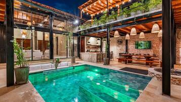 A modern outdoor pool area with a glass enclosure, a dining area with a table and chairs, and a kitchen visible through the glass.