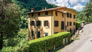 A yellow building with a tiled roof, multiple windows with shutters, and a curved road nearby. - Apartment in Villa Poesia - Bagni di Lucca (Bagni di Lucca)
