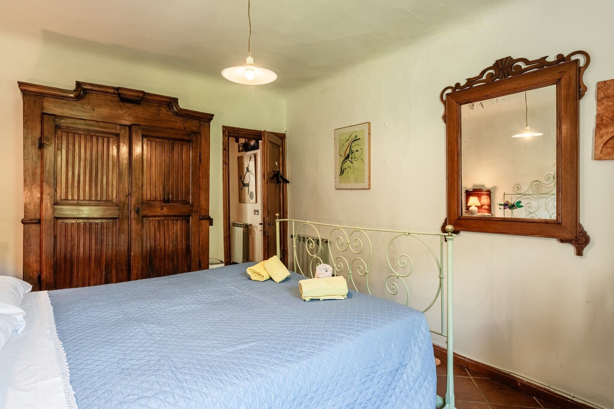 A bedroom with a bed, wooden wardrobe, a mirror, and a framed artwork on the wall.
