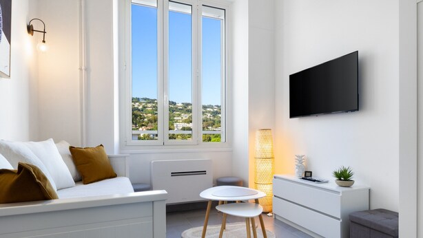 Een moderne woonkamer met een bank, een aan de muur bevestigde televisie, een klein tafeltje en een stadsgezicht door grote ramen.