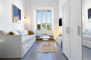 Ein schmaler Raum mit Bett, kleinem Tisch, Stuhl, Fenster mit Blick und Wand-Fernseher.