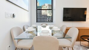 A dining area with a glass table, four chairs, and a window with a city view. - Urban Escape : Bright 2BR/2BA in FiDi (New York)