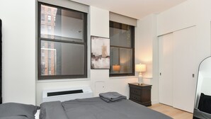 A bedroom with a bed, a bedside table, a lamp, a mirror, and a window with a view of buildings. - Urban Escape : Bright 2BR/2BA in FiDi (New York)