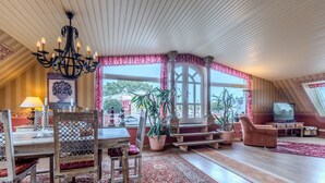 Amplia sala de estar con una mesa de comedor de madera, un candelabro, un ventanal con cortinas, un televisor y una planta en maceta.