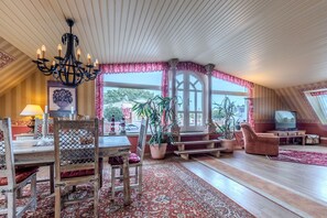 A spacious living area with a wooden dining table, a chandelier, a large window with curtains, a television, and a potted plant. - Apartment Meerblick 17 with Sea View and Private Rooftop Terrace (Binz)