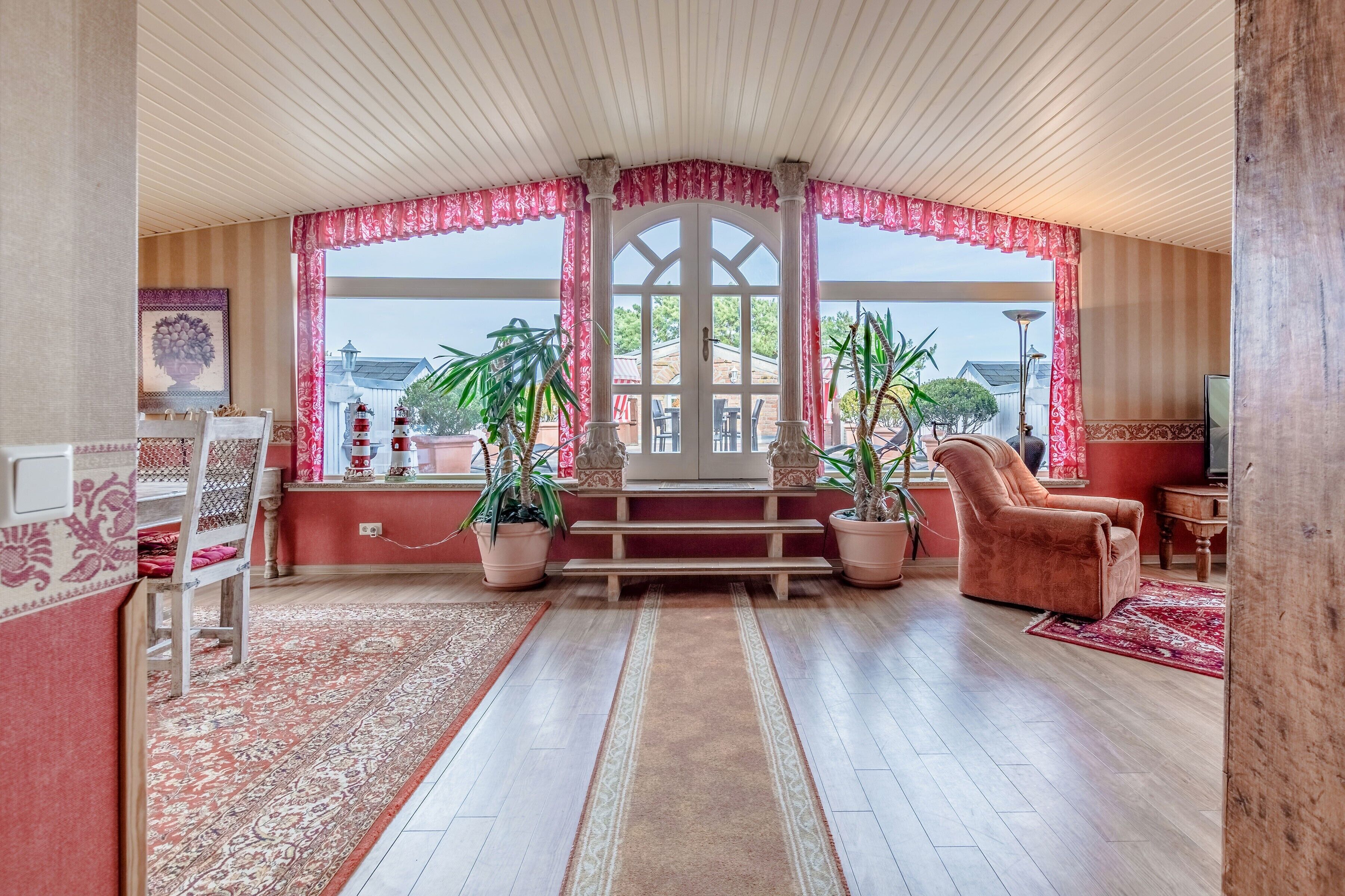 Ein Zimmer mit Holzboden, einem rot-weißen Teppich, einer hölzernen Bank, einem rosa Stuhl und Topfpflanzen.